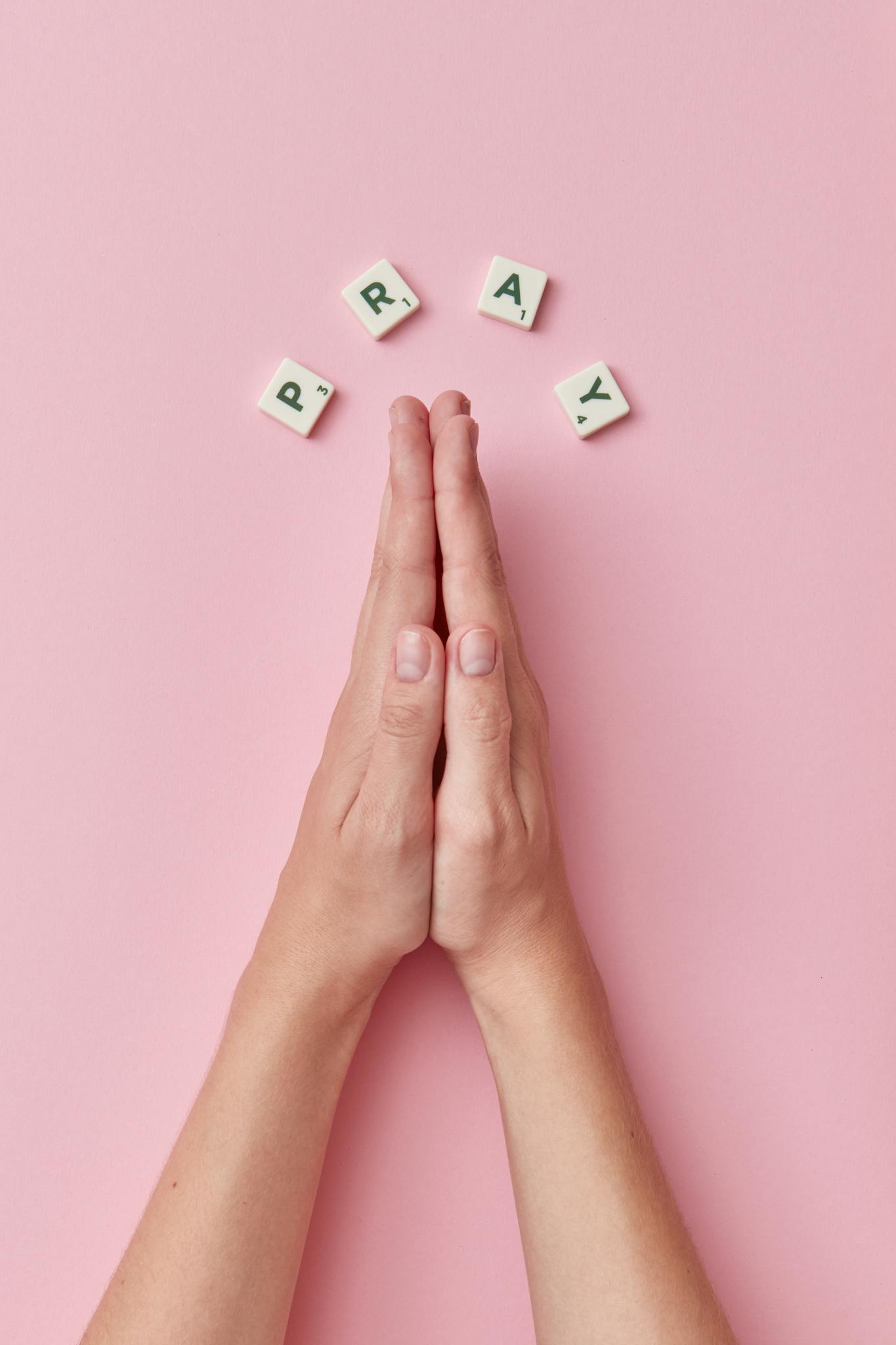 Top view of praying hands with Scrabble letters spelling 'PRAY' on a pink background.